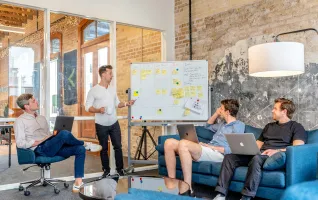 Drei Männer sitzen am Laptop und beobachten den Mann neben dem Whiteboard bei der Mitarbeiterschulung