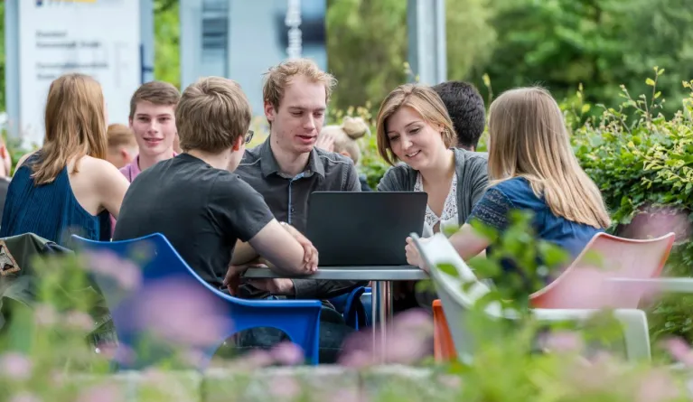 Studierende am Campus in Stade sitzen um einen runden Tisch herum draußen am Campus in Stade und schauen gemeinsam auf einen Laptop.