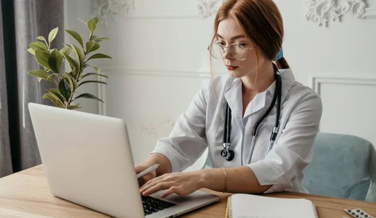 woman doctor wearing white coat and stethoscope typing on laptop