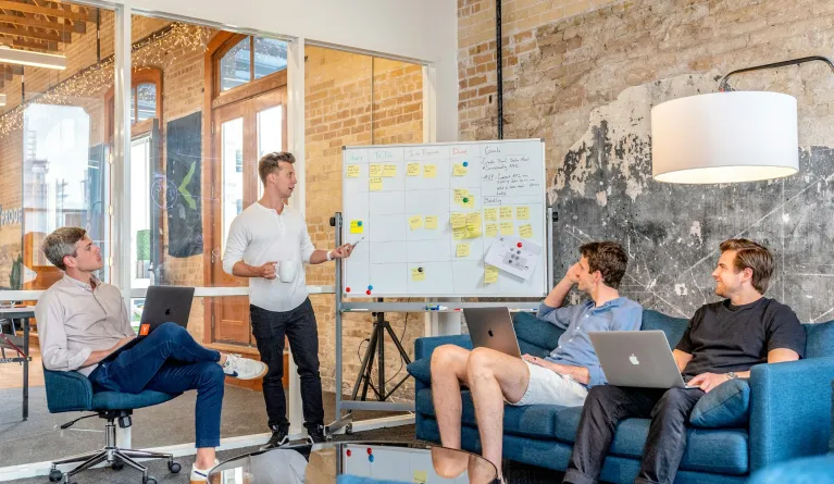 Drei Männer sitzen am Laptop und beobachten den Mann neben dem Whiteboard bei der Mitarbeiterschulung